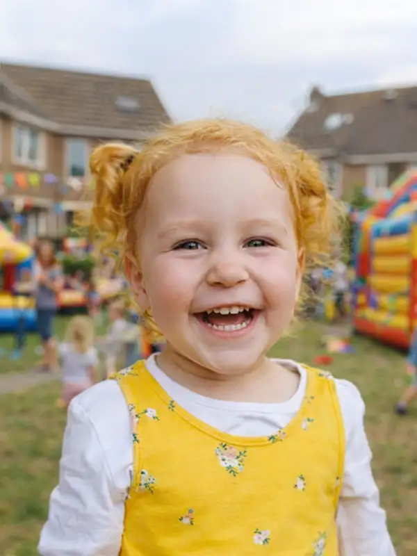 Blij lachend meisje in gele jurk op een buurtfeest in een Nederlandse woonwijk, met kleurrijke springkussens en spelende kinderen op de achtergrond.